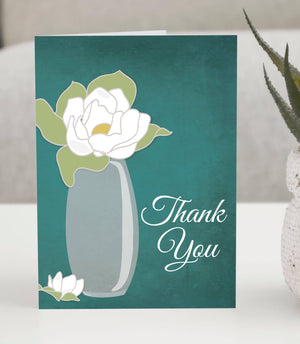 Thank You greeting card featuring a white magnolia flower in a glass vase on a rich teal background with elegant script text.