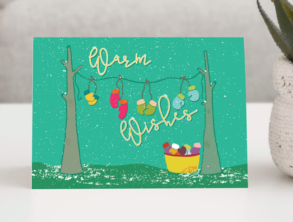 Festive holiday greeting card featuring a charming winter scene with colorful mittens hanging from a clothesline between two trees. 
