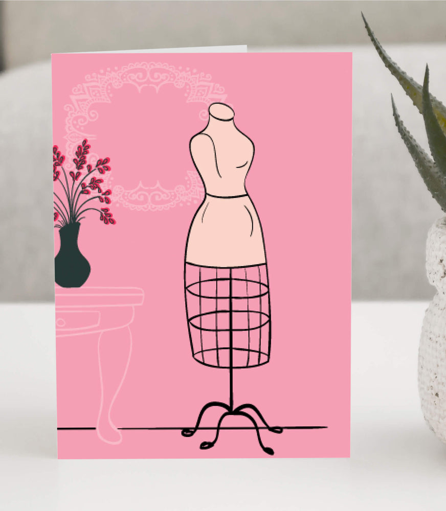 Greeting card featuring a vintage dress form with a wire skirt on a pink background, accented with a decorative lace frame and floral vase.  